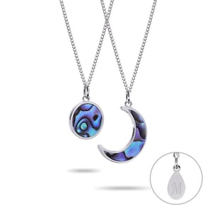 Puede incluir: Dos collares plateados con colgantes. Uno es un círculo con un diseño de concha de abulón azul y morado. El otro es una media luna con un diseño similar. También es visible un pequeño dije en forma de lágrima con la letra "M".
