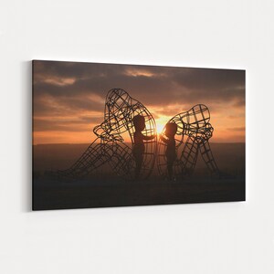 May include: Silhouette of two children standing inside a large metal sculpture of two adults, set against a sunset sky.
