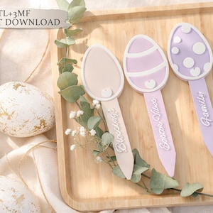 May include: Three pastel-coloured Easter egg-shaped garden markers. Each marker has a different design and the words "Blessings", "Easter Vibes", and "Family". The markers are on a wooden tray with decorative eggs. The text "STL+3MF INSTANT DOWNLOAD" is visible.