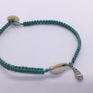 Op de afbeelding: Een geknoopt armband van tealblauw koord met een witte schelpbedel en een zilveren schelpbedel.