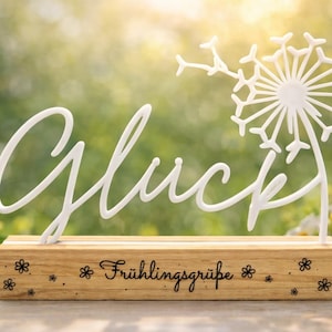 Könnte beinhalten: Ein weißes Schild mit dem Wort "Glück" in Schreibschrift und einem Pusteblumen-Design, montiert auf einem Holzsockel mit dem Text "Frühlingsgrüße". Gelbe Narzissen und weiße Gänseblümchen im Hintergrund.