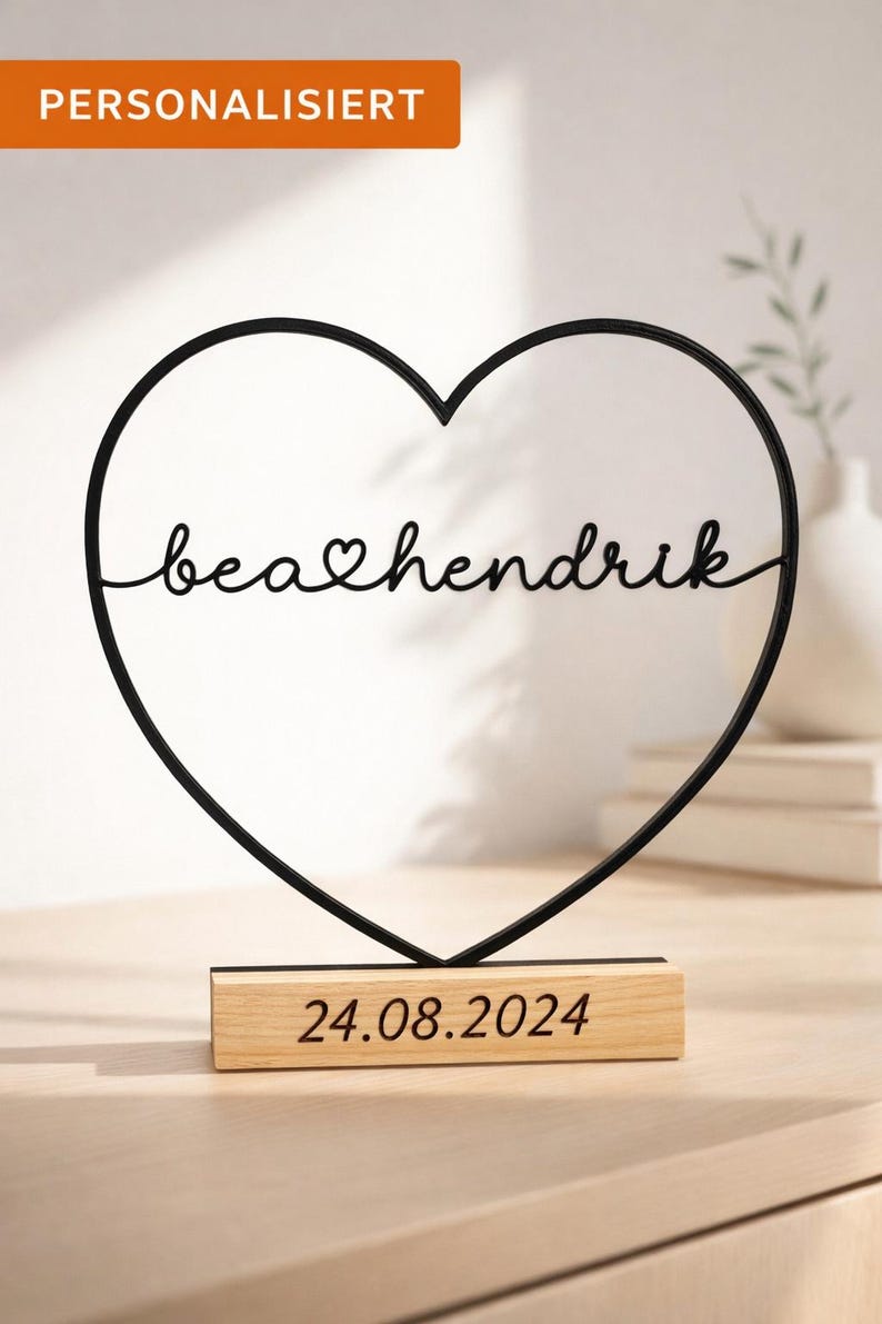 May include: A black metal heart-shaped wire sculpture featuring the cursive text "bea" followed by a small heart symbol and "hendrik" in the center. The sculpture is mounted on a rectangular light wood base with the date "24.08.2024" printed on it.
