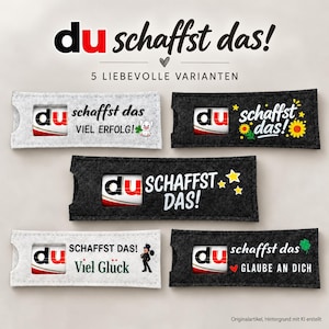 Könnte beinhalten: Fünf rechteckige Filzhüllen mit Sichtfenstern, in denen Schokoriegel stecken. Der Text lautet du schaffst das! mit verschiedenen deutschen Phrasen wie Viel Erfolg, Viel Glück und Glaube an dich, ergänzt durch Symbole wie Schwein, Sonnenblumen, Sterne und Schornsteinfeger.