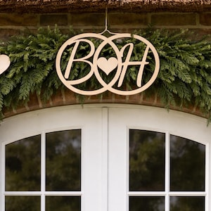Könnte beinhalten: Holzhochzeitsdekoration mit ineinandergreifenden Kreisen mit den Buchstaben "B" und "H" und einem Herz. Sie hängt über einer weißen Tür, geschmückt mit einer grünen Girlande und Holzherz-Ornamenten.