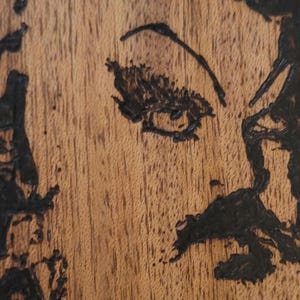 May include: A close-up of a wood-burned portrait of a person with a moustache. The portrait is on a piece of wood with a natural grain.
