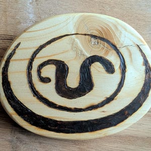 May include: A wooden circle with a black and brown design of a stylized letter U. The design is surrounded by a black circle.