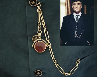 May include: A gold-colored pocket watch chain with a red circular pendant and decorative links, attached to a dark green suit jacket. The jacket has matching buttons with a starburst design. A photo of a man in a suit is in the background.