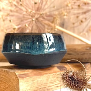 Könnte beinhalten: Eine Keramikschale mit einem glänzend tiefblauen glasierten oberen Teil und einer matten, texturierten schwarzen Basis. Die Schale hat einen breiten, abgerundeten Körper, der sich nach unten verjüngt, und zeigt einen deutlichen Kontrast zwischen der glatten blauen Oberfläche und dem dunklen, körnigen unteren Bereich.