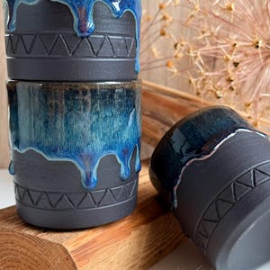 Könnte beinhalten: Drei Keramikbecher mit einer mattschwarzen Basis und einem eingravierten geometrischen Dreiecksmuster. Der obere Teil jedes Bechers ist mit einer glänzenden, tiefblauen Glasur versehen, die in einem vertikalen, fließenden Muster herabläuft.