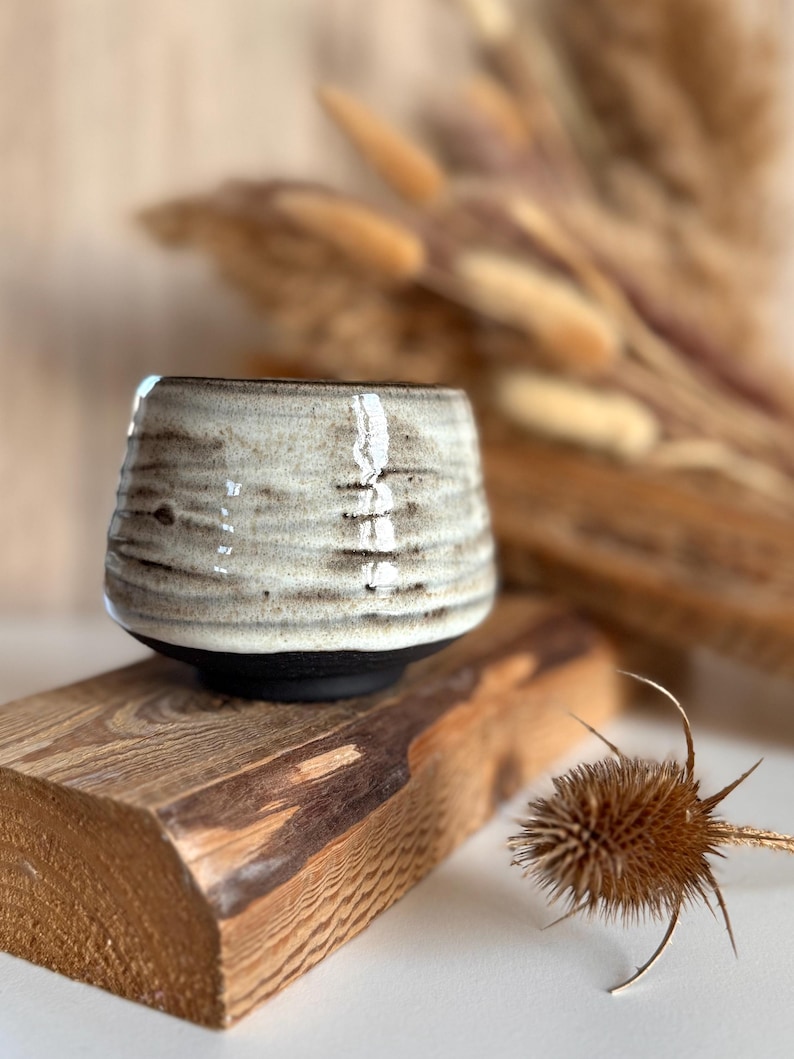 K&ouml;nnte beinhalten: Eine kleine Keramiktasse mit abgerundeter Form, die eine texturierte cremewei&szlig;e Glasur mit horizontalen braunen Streifen im oberen Bereich und einen soliden mattschwarzen Boden aufweist. Die Tasse hat eine weite &Ouml;ffnung und einen dicken Rand.