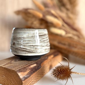 K&ouml;nnte beinhalten: Ein Keramikbecher mit einem texturierten, cremewei&szlig; glasierten Oberteil, das horizontale Rillen und braune Sprenkel aufweist, im Kontrast zu einem glatten, mattschwarzen Boden. Das Gef&auml;&szlig; hat eine breite, abgerundete Form und einen dicken Rand.
