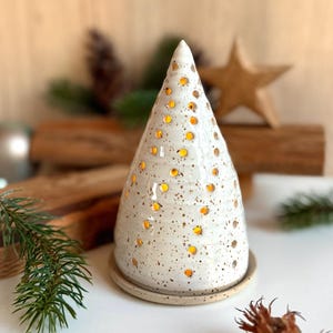 Könnte beinhalten: Eine konische Keramikskulptur in Baumform mit weiß glasierter, gesprenkelter Oberfläche. Das Objekt weist zahlreiche kleine kreisförmige Aussparungen auf, durch die ein warmes gelbes Licht aus dem Inneren scheint, auf einem runden Sockel.