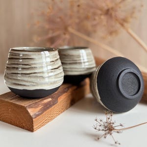 K&ouml;nnte beinhalten: Drei Keramikbecher mit zweifarbigem Design, bestehend aus einem strukturierten, cremefarbenen Oberteil und einem mattschwarzen Boden. Die Becher haben eine bauchige Form mit horizontalen Rillen und einem kreisf&ouml;rmigen Spiralmuster am Boden.