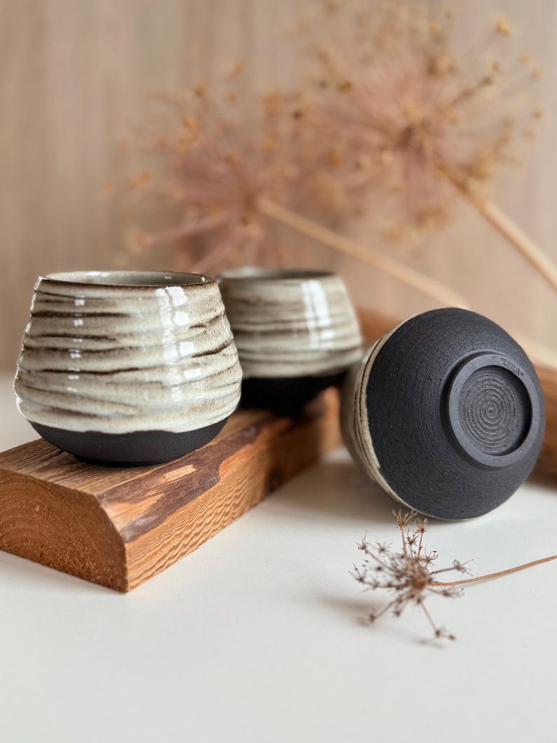 K&ouml;nnte beinhalten: Drei Keramikbecher mit zweifarbigem Design, bestehend aus einem strukturierten, cremefarbenen Oberteil und einem mattschwarzen Boden. Die Becher haben eine bauchige Form mit horizontalen Rillen und einem kreisf&ouml;rmigen Spiralmuster am Boden.