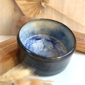 Könnte beinhalten: Eine kleine, runde Keramikschale mit einer glatten, dunklen Außenseite und einer reaktiven Glasur im Inneren. Das Innere zeigt ein tiefblaues Zentrum, das in einen helleren, gesprenkelten blau-cremefarbenen Rand übergeht und einen Kontrast bildet.