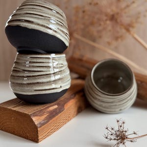 K&ouml;nnte beinhalten: Drei Keramikbecher mit abgerundeter Form, einer mattschwarzen Basis und einer texturierten, cremefarbenen Glasur im oberen Bereich. Die Becher weisen horizontale Rillen im glasierten Teil auf, wobei ein Becher auf einem anderen steht und einer daneben liegt.
