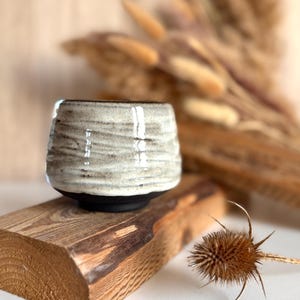 K&ouml;nnte beinhalten: Eine kleine, runde Keramiktasse mit einem texturierten, cremewei&szlig;en glasierten Oberteil mit horizontalen Rillen und einem glatten, mattschwarzen Boden. Die Tasse hat eine weite &Ouml;ffnung und einen verj&uuml;ngten Boden, der den Kontrast der Oberfl&auml;chen betont.