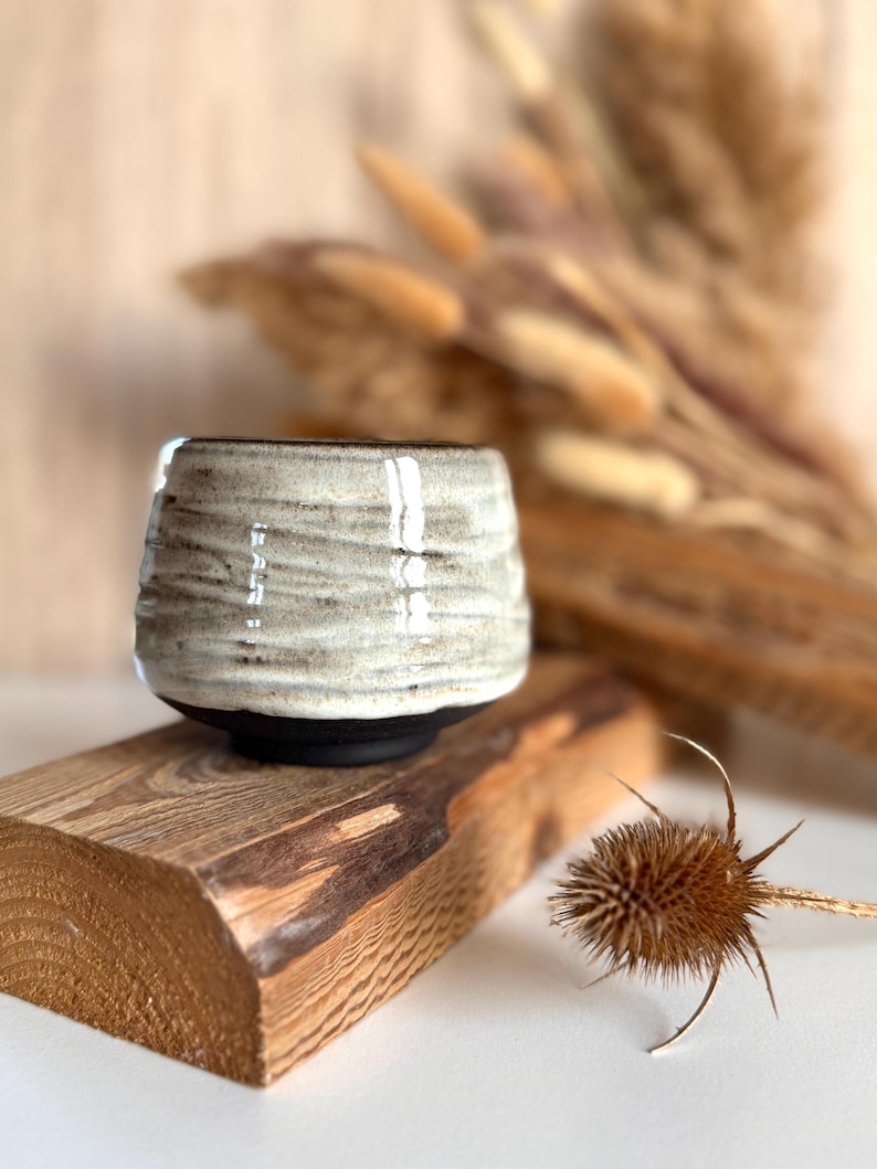 K&ouml;nnte beinhalten: Eine kleine, runde Keramiktasse mit einem texturierten, cremewei&szlig;en glasierten Oberteil mit horizontalen Rillen und einem glatten, mattschwarzen Boden. Die Tasse hat eine weite &Ouml;ffnung und einen verj&uuml;ngten Boden, der den Kontrast der Oberfl&auml;chen betont.