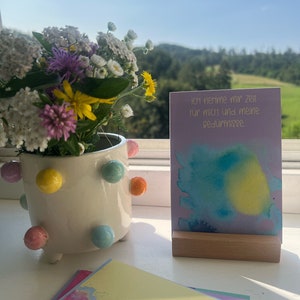 May include: A purple card with a watercolor background and the text "Ich nehme mir Zeit f&uuml;r mich und meine Bed&uuml;rfnisse."  The card is sitting on a wooden stand. A bouquet of wildflowers is in a white ceramic pot with colorful polka dots.