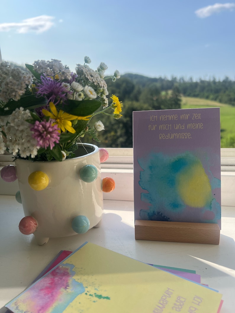 May include: A purple card with a watercolor background and the text "Ich nehme mir Zeit f&uuml;r mich und meine Bed&uuml;rfnisse."  The card is sitting on a wooden stand. A bouquet of wildflowers is in a white ceramic pot with colorful polka dots.