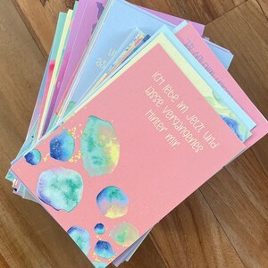 May include: A stack of pink, blue, and green cards with watercolor designs. The top card has the text "ich lebe im jetzt und lasse vergangenes hinter mir."