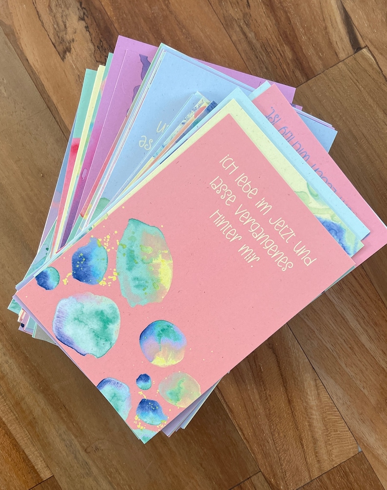 May include: A stack of pink, blue, and green cards with watercolor designs. The top card has the text "ich lebe im jetzt und lasse vergangenes hinter mir."