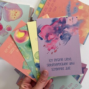 May include: A stack of colorful cards with watercolor designs and inspirational quotes in German. The top card reads "Ich strahle Liebe, Selbstvertrauen und Sch&ouml;nheit aus."