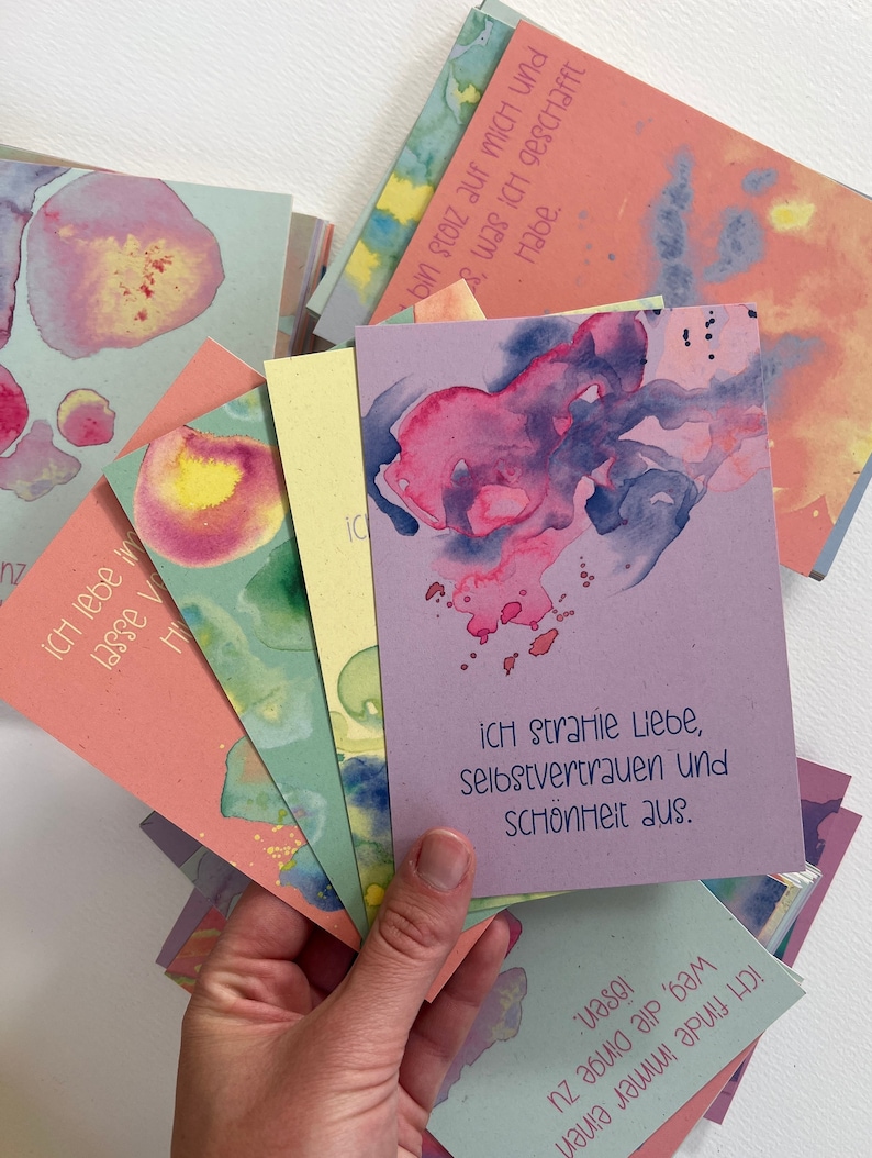 May include: A stack of colorful cards with watercolor designs and inspirational quotes in German. The top card reads "Ich strahle Liebe, Selbstvertrauen und Sch&ouml;nheit aus."