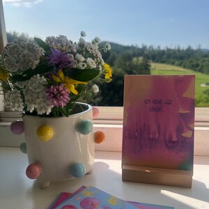 May include: A set of colorful watercolor cards with the text "ICH liebe das Leben" on one card. The cards are displayed on a wooden stand and a white ceramic pot with colorful polka dots.