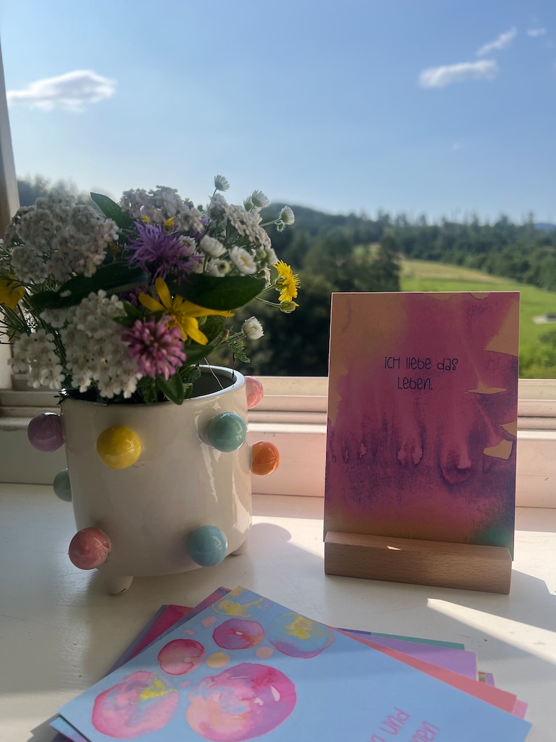 May include: A set of colorful watercolor cards with the text "ICH liebe das Leben" on one card. The cards are displayed on a wooden stand and a white ceramic pot with colorful polka dots.