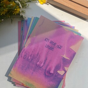 May include: A stack of colorful cards with a watercolor background. The top card has the text "Ich liebe das Leben" written in black.