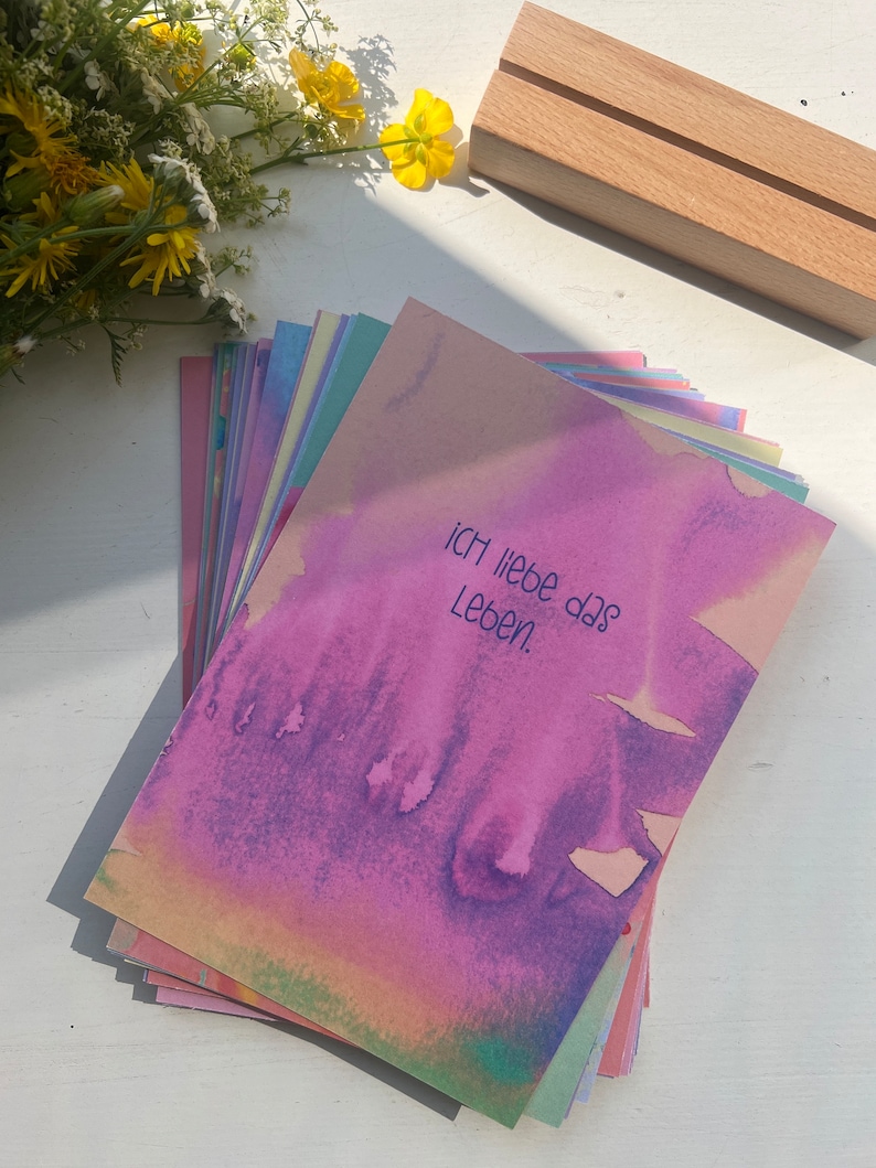 May include: A stack of colorful cards with a watercolor background. The top card has the text "Ich liebe das Leben" written in black.