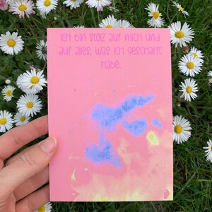 May include: A pink card with a blue and yellow watercolor design. The card has the text "Ich bin stolz auf mich und auf alles, was ich geschafft habe." written in black ink.