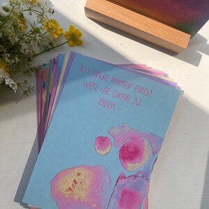 May include: A stack of blue, pink, and yellow affirmation cards with the text "Ich finde immer einen Weg die Dinge zu l&ouml;sen."  The cards have watercolor-like designs with pink, yellow, and blue colors.