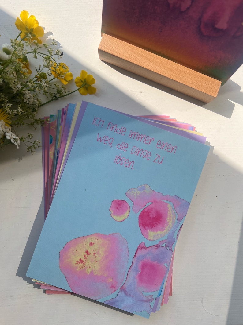 May include: A stack of blue, pink, and yellow affirmation cards with the text "Ich finde immer einen Weg die Dinge zu l&ouml;sen."  The cards have watercolor-like designs with pink, yellow, and blue colors.