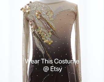 May include: A figure skating costume with a nude mesh bodice and long sleeves. The bodice is embellished with sequins and rhinestones in a gradient pattern from light to dark. The skirt is a dark brown with a layered tulle overlay. The text "Wear This Costume @ Etsy" is printed on the skirt.