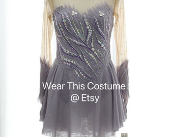 May include: A long-sleeved figure skating dress with a purple and silver design. The dress has a sheer bodice and a full skirt. The dress is decorated with rhinestones and beads. "Wear This Costume @ Etsy" is written on the skirt.