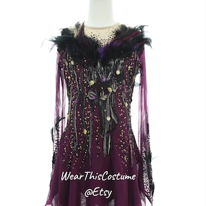 Pode incluir: Um vestido de patinação artística de cor bordô com mangas compridas e acabamento de penas pretas. O vestido é adornado com strass e lantejoulas pretas, douradas e roxas. A saia tem uma bainha irregular. "WearThisCostume @Etsy" está impresso no vestido.