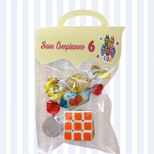 Può includere: Un sacchetto trasparente di cellophane pieno di caramelle incartate e un Cubo di Rubik. La borsa ha un manico giallo e una parte superiore decorativa con la scritta "Buon Compleanno 6" e una grafica a cartoni animati. Le caramelle sono di vari colori.