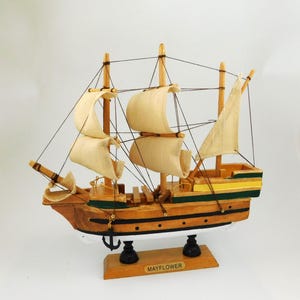 May include: A wooden model of the Mayflower ship with beige sails and a brown hull. The ship has black rigging and is mounted on a wooden base. The base has the word "MAYFLOWER" printed on it.