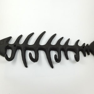 May include: A black cast iron wall hook rack shaped like a fish skeleton. The horizontal design features four curved hooks extending downwards from the spine, with two visible mounting holes positioned along the central bone structure.
