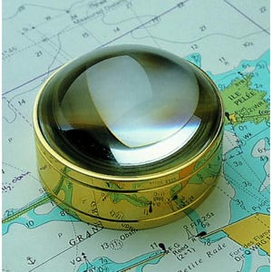 May include: A brass magnifying glass with a clear, rounded lens. The magnifier rests on a nautical chart, with visible text and lines. The lens reflects light, highlighting its convex shape. A tool for navigation and map reading.