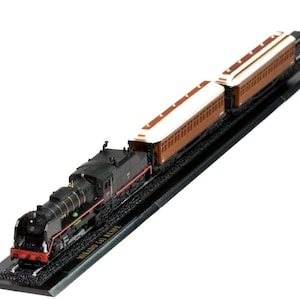 May include: A model train set with a black steam locomotive, red and gold detailing, and two brown and white passenger carriages. The model sits on a black track base, with the text "MIKADO 141 RENFE" visible.