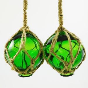 May include: Two spherical green glass floats encased in a decorative tan jute rope netting. Each glass orb features a thick, braided jute rope loop at the top for hanging, creating a nautical aesthetic with the vibrant, translucent green glass.
