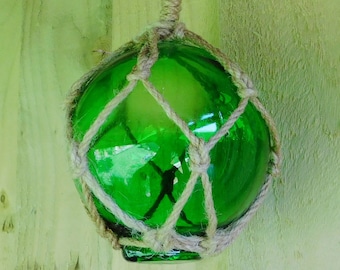 May include: A green glass float, spherical in shape, encased in a natural rope netting. A thicker rope is attached for hanging. The glass has a shiny surface, and the rope is a light brown colour. The background is a light green wooden surface.