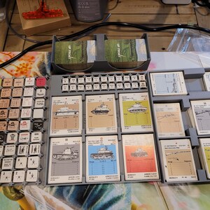 May include: A tabletop war game setup with various components. Includes a keyboard-like tile grid with small square tiles, a card holder with cards labeled "UP FRONT", and multiple trays holding illustrated cards depicting tanks and other military equipment.
