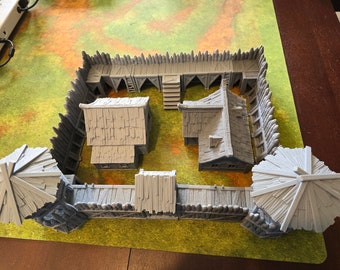 Puede incluir: Un modelo 3D impreso en gris de un pueblo medieval con una valla de madera, dos casas y dos torres. El modelo está colocado sobre una superficie verde de hierba.