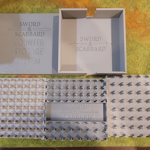 以下が含まれることがあります： 蓋と容器に「SWORD & SCABBARD」の文字が入った、明るい灰色のカウンター収納システム。システムには、長方形の容器、蓋、およびいくつかのインターロッキンググリッドピースが含まれています。これらの部品は、緑と黄色の表面に配置されています。