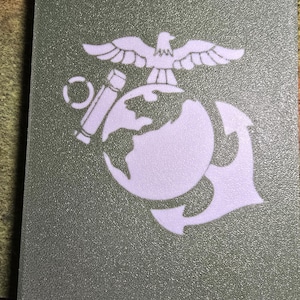 May include: A rectangular, olive green card featuring a white emblem of an eagle atop a globe, anchor, and a cylindrical object. The emblem is a symbol of the United States Marine Corps. The card has a textured surface.