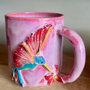 Puede incluir: Una taza de cerámica rosa con un diseño de colibrí esculpido. El colibrí tiene detalles rojos, azules y amarillos. La taza tiene un acabado brillante y un asa curva. El colibrí está representado cerca de una flor roja.